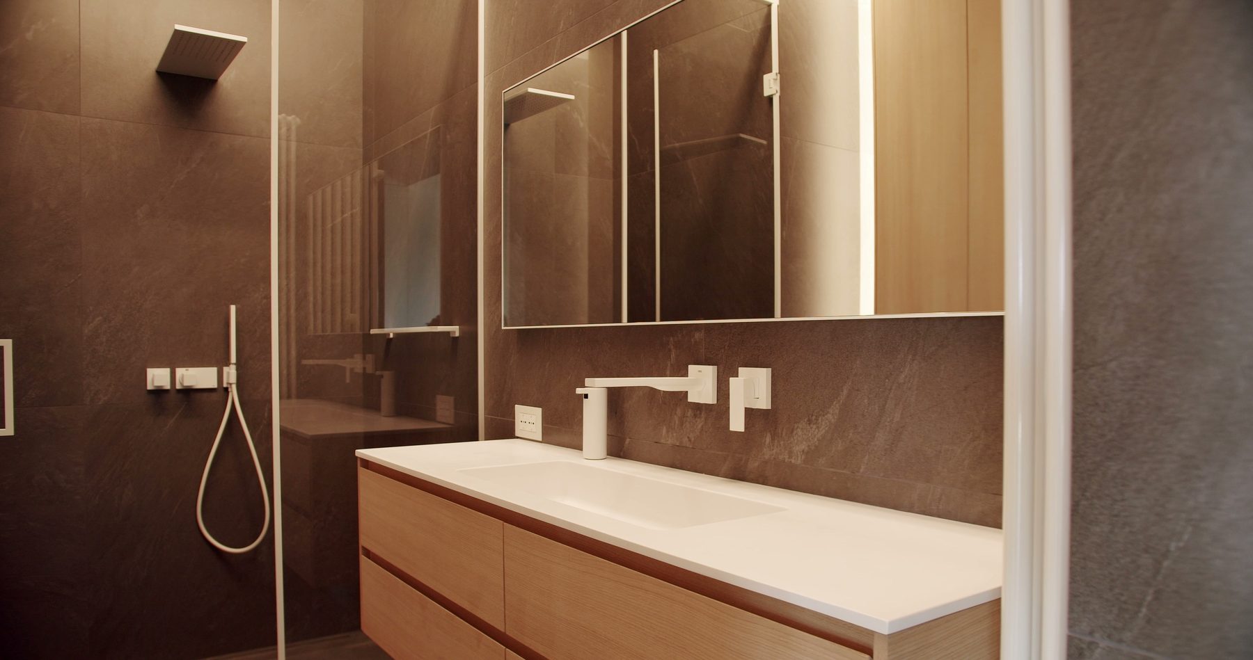 A floating wood vanity with brass fixtures beside a stone-tiled shower